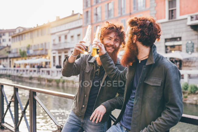 Jovens gêmeos hipster masculinos com cabelos vermelhos e barbas fazendo um brinde na orla do canal — Fotografia de Stock