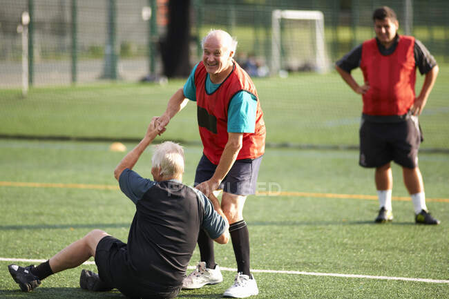 Senior man helping friend stand up on football pitch — Stock Photo