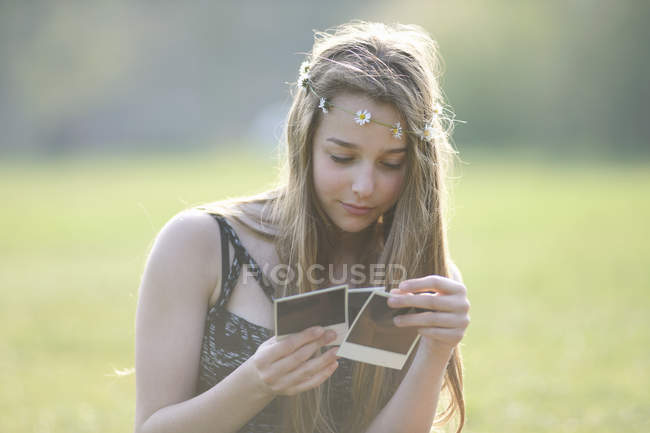 Adolescente usando coberturas para a cabeça cadeia margarida olhando para fotografias instantâneas no parque — Fotografia de Stock