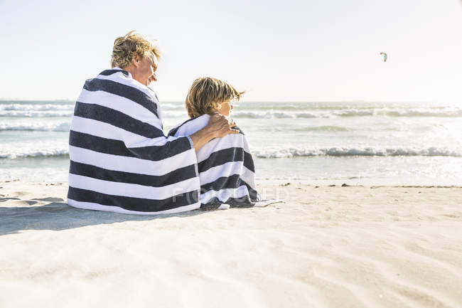 Vista trasera del padre y el hijo sentados en la playa envueltos en manta - foto de stock