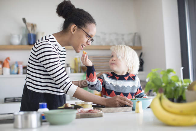Filho alimentando mãe comida na cozinha em casa — Fotografia de Stock