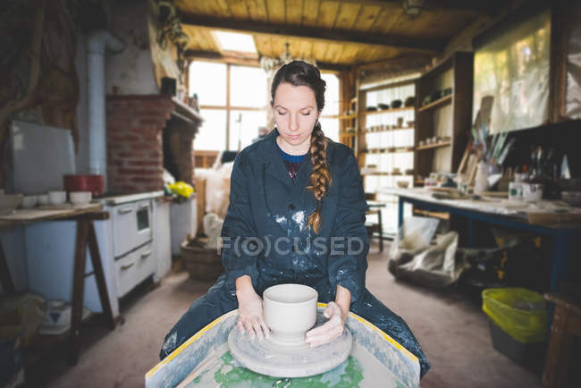 Vista frontal da jovem mulher na oficina sentada na roda de cerâmica fazendo panela de barro, olhando para baixo — Fotografia de Stock