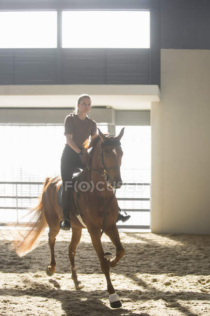 Woman trotting on chestnut horse in indoor paddock — Stock Photo