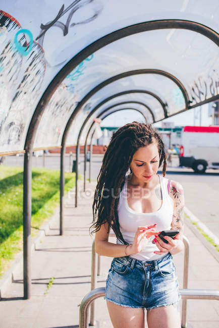 Молодая женщина с дредами с сенсорным экраном смартфона в городском автобусном приюте — стоковое фото
