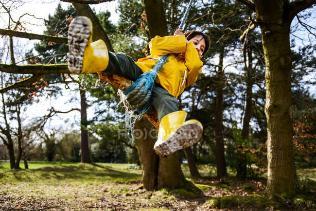 Niño en anorak amarillo balanceándose del árbol del parque - foto de stock