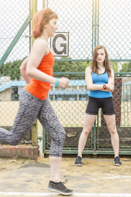 Runner passando jovem mulher de pé ao lado de campo de esportes, Londres, Reino Unido — Fotografia de Stock