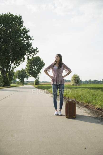 Jovem mulher de pé na estrada de campo com mala — Fotografia de Stock