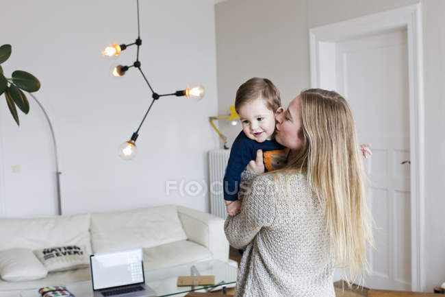 Mulher adulta média beijando bebê filho na bochecha na sala de estar — Fotografia de Stock