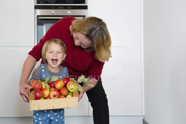 Porträt eines weiblichen Kleinkindes und einer Mutter, die in der Küche eine Kiste mit Äpfeln hält — Stockfoto