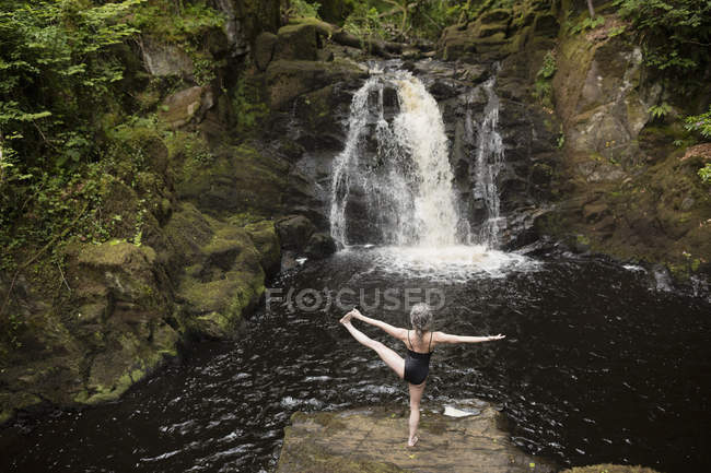 Rückansicht einer reifen Frau, die vor dem Wasserfall Yoga praktiziert — Stockfoto