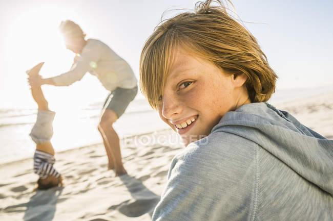 Хлопчик на пляжі з батьком і братом дивиться через плече на камеру посміхаючись — стокове фото