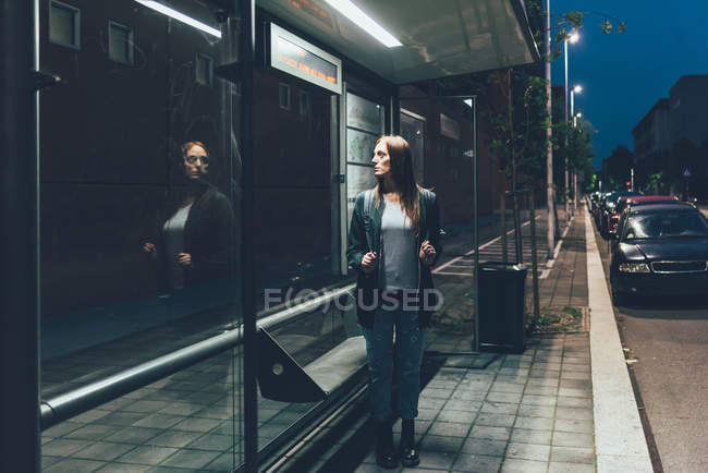 Jeune femme regardant la réflexion en attendant à l'arrêt de bus la nuit — Photo de stock