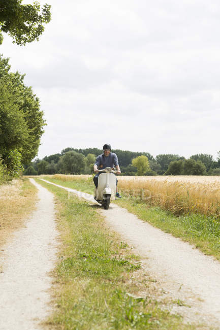 Зрелый мужчина едет на мотороллере по грунтовой дорожке с дочерью, держащейся за талию — стоковое фото