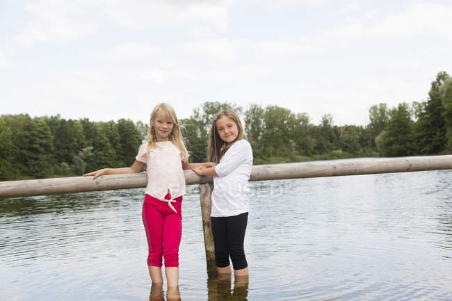 Portrait of two girls holding onto fence whilst paddling in rural lake — Stock Photo