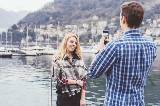 Jeune homme sur le front de mer photographiant petite amie, Lac de Côme, Italie — Photo de stock