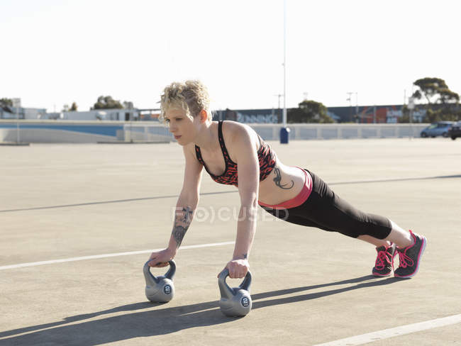 Young woman doing push ups on kettlebells in parking lot — Stock Photo