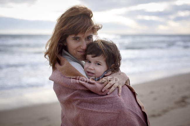 Madre e figlia avvolto in coperta abbracciare sulla spiaggia — Foto stock