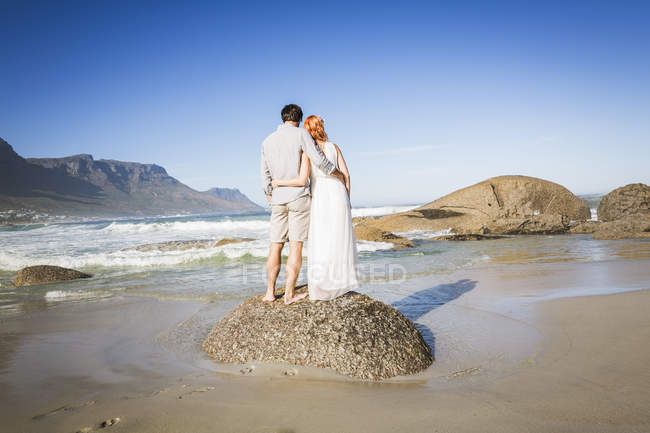 Повна довжина вигляд ззаду пари, руки навколо кожного, стоячи на скелі на березі — стокове фото
