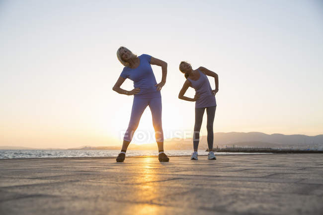 Two cheerful women stretching by beach at sunset — Stock Photo