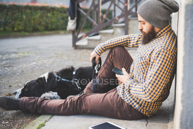 Homem adulto médio sentado ao ar livre com cão, usando smartphone — Fotografia de Stock