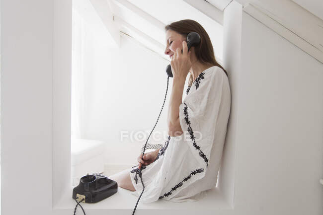 Vista lateral de una mujer madura sentada apoyada contra la pared usando un teléfono fijo sonriendo - foto de stock