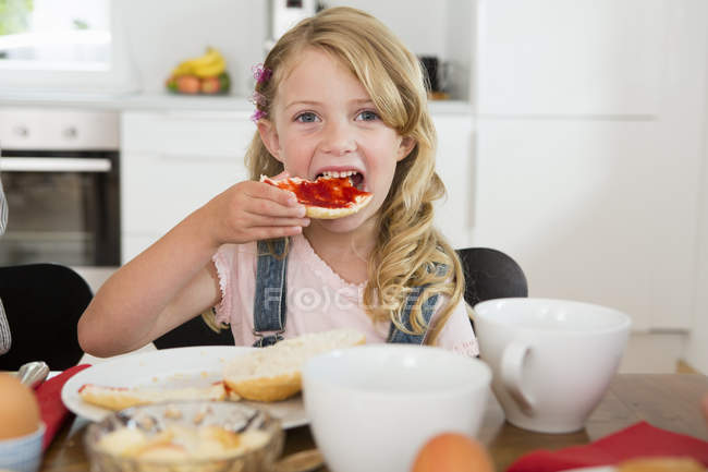 Girl eating toast with jam at kitchen table and looking in camera — Stock Photo