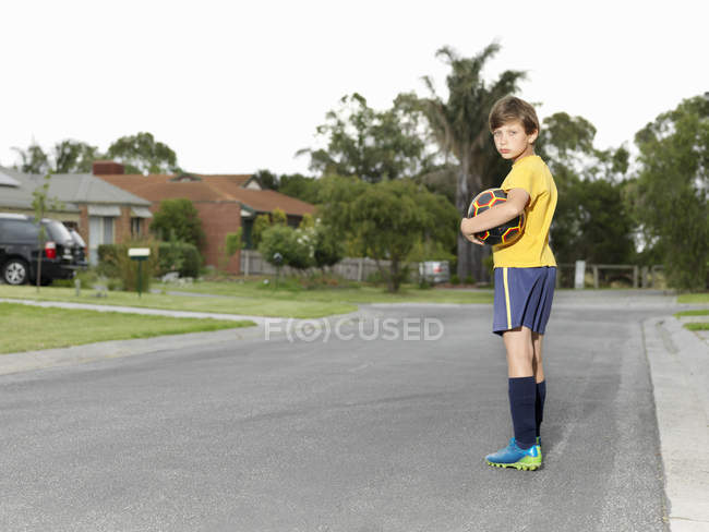 Portrait d'un garçon maussade tenant un ballon de football sur une route de banlieue — Photo de stock