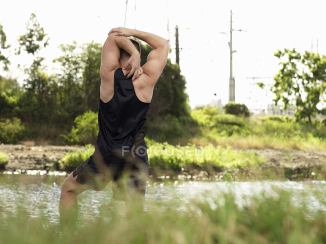 Молодой человек тренируется у реки, вытягивает руки, смотрит сзади — стоковое фото
