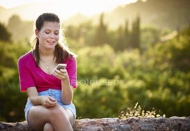 Teenager-Mädchen sitzt auf Steinmauer und schaut auf Smartphone, Mallorca, Spanien — Stockfoto