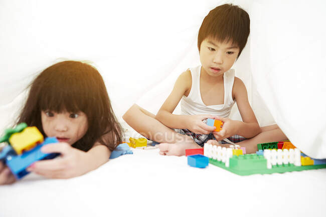 Junge chinesische Jungen und Mädchen spielen im Bett mit ihren Spielzeugen unter den Bettlaken — Stockfoto