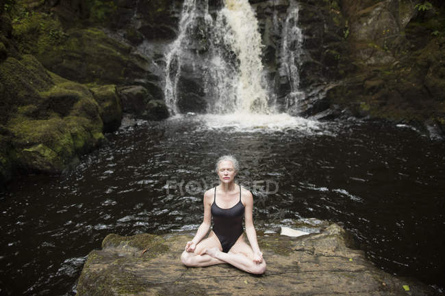 Mature woman practicing lotus pose in front of waterfall — Stock Photo