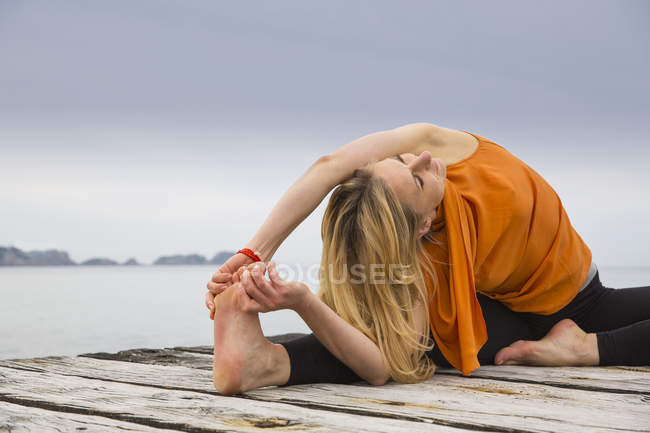 Середня доросла жінка торкається пальців, практикуючи йогу на дерев'яному морському пірсі — стокове фото