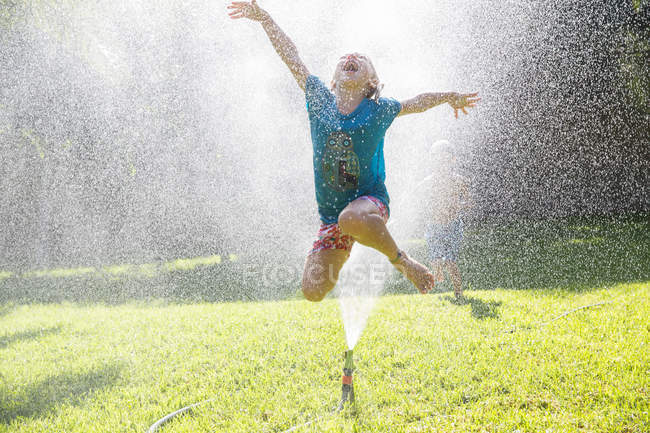 Chica saltando sobre el aspersor de agua en el jardín - foto de stock