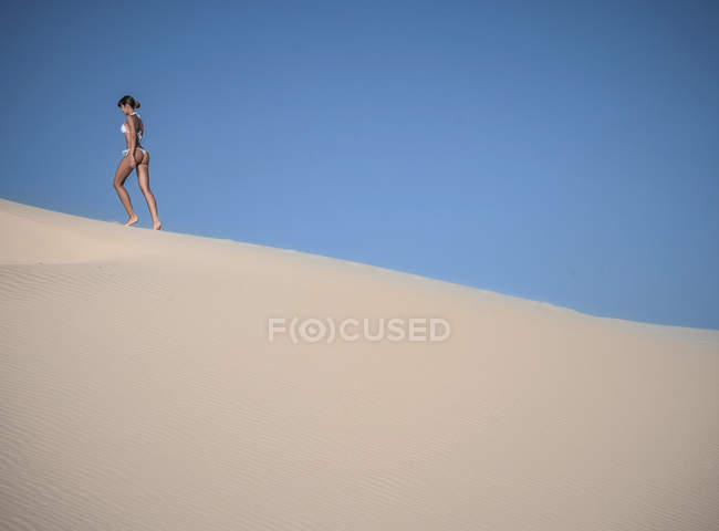 Jeune femme marchant sur une dune de sable contre un ciel bleu clair — Photo de stock