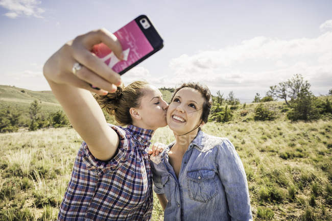 Две молодые женщины делают селфи со смартфоном с поцелуем в щеку, Бриджер, Монтана, США — стоковое фото