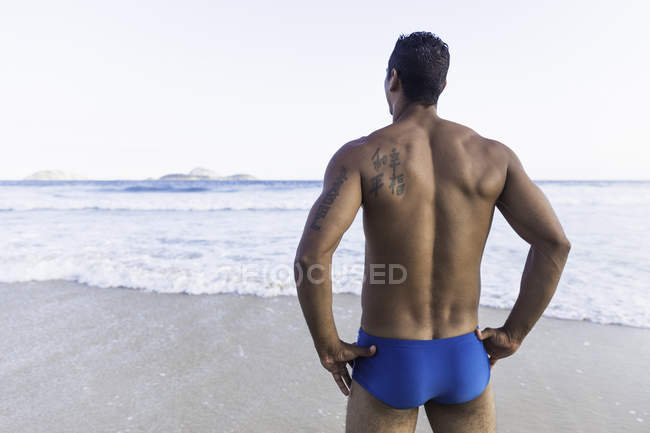 Mid adult man wearing swimming trunks, looking out to sea, rear view — Stock Photo