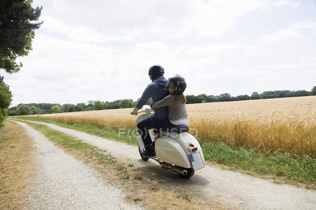 Vista trasera de un hombre maduro y una hija montando un scooter a lo largo de la pista de tierra - foto de stock