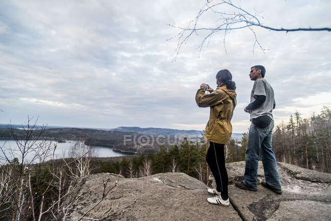 Jeune couple au sommet de la formation rocheuse photographiant paysage — Photo de stock