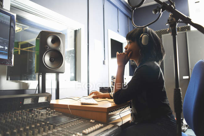 Side view of young woman wearing headphones, sitting at mixing desk in recording studio looking at monitor — Stock Photo