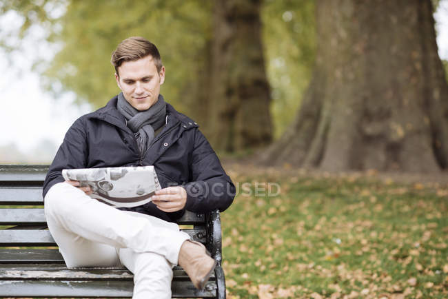 Молодой человек читает газету на скамейке в парке — стоковое фото