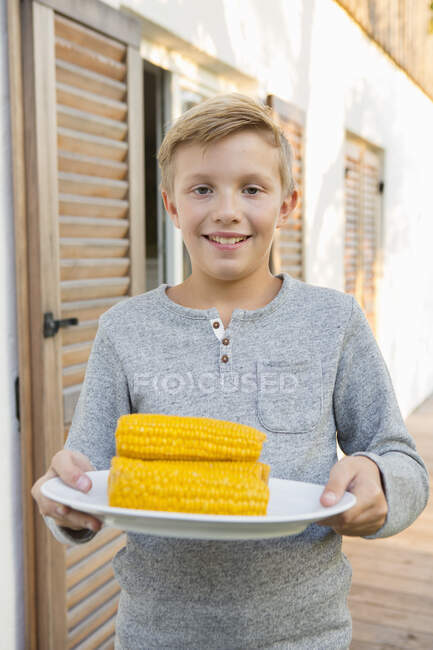 Porträt eines Jungen, der einen Teller mit Maiskolben zum Grillen im Garten trägt — Stockfoto