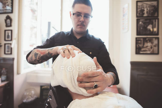 Barbeiro envolvendo toalha em torno do rosto do cliente na barbearia — Fotografia de Stock