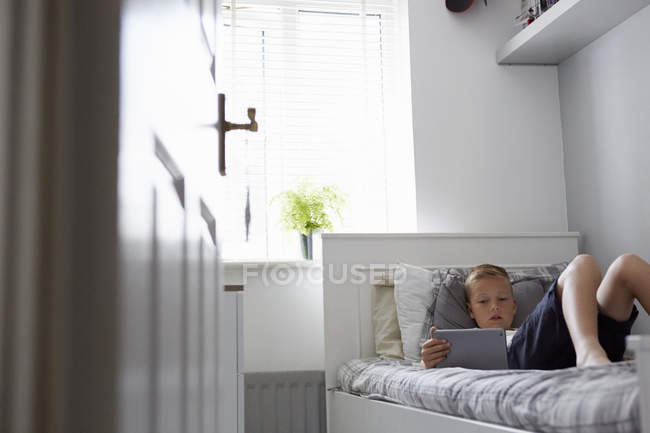 Vue à travers la porte du garçon allongé sur le lit regardant vers le bas à la tablette numérique — Photo de stock