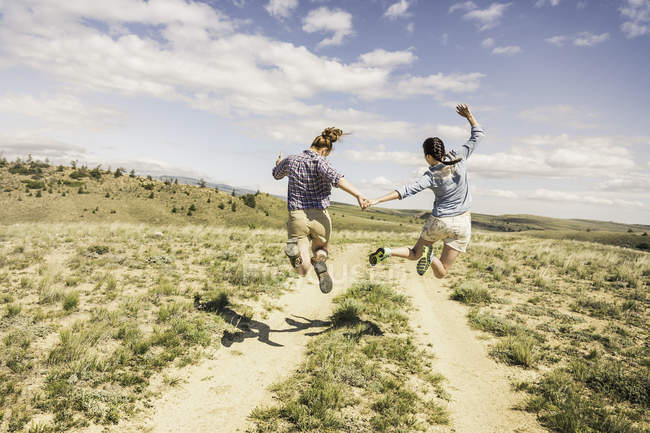Rückansicht von zwei jungen Frauen, die mitten in der Luft über Feldweg, Brücke, Montana, USA springen — Stockfoto