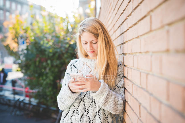 Mujer mensajes de texto en el teléfono inteligente, contra la pared de ladrillo - foto de stock