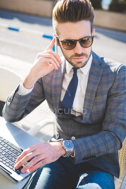 Stylish young businessman using smartphone and laptop at sidewalk cafe — Stock Photo