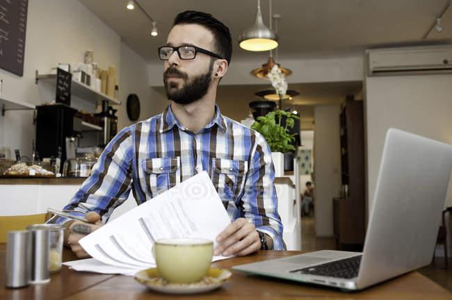 Männlicher Kunde liest Papierkram und benutzt Laptop am Cafétisch — Stockfoto