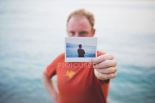 Man holding up photograph of himself by lake — Stock Photo