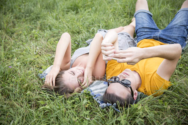 Молода пара лежить на траві в полі, дивлячись на смартфон — стокове фото