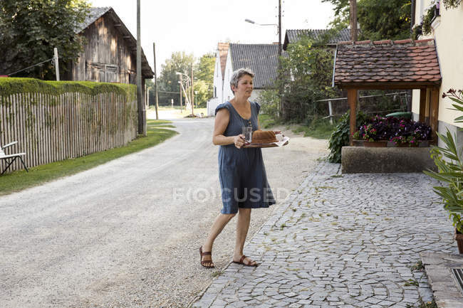 Donna matura attraverso il percorso, vassoio di trasporto con torta e bevande — Foto stock
